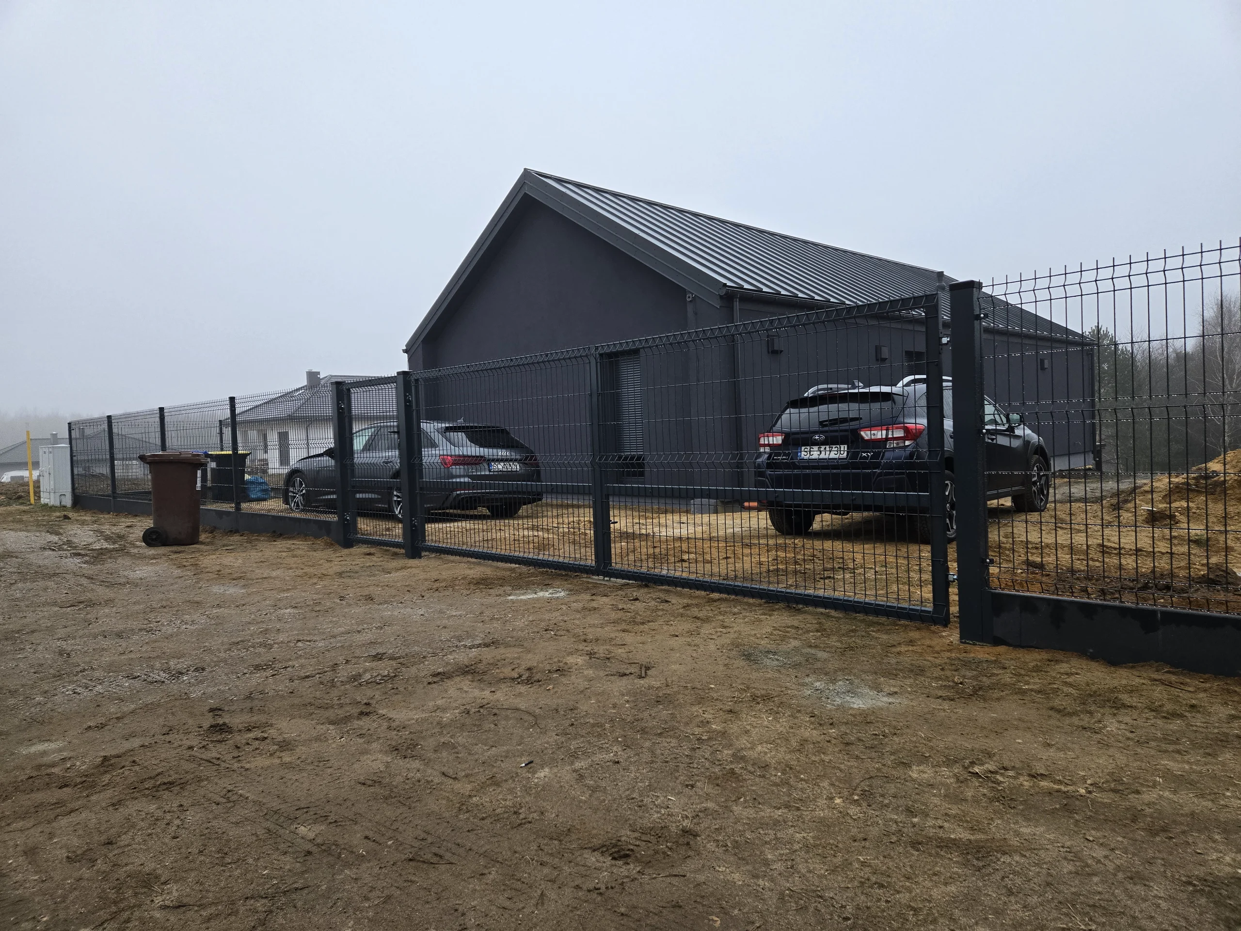 Ogrodzenie panelowe z bramą przesuwaną przy nowoczesnym ciemnym domu i zaparkowanych samochodach